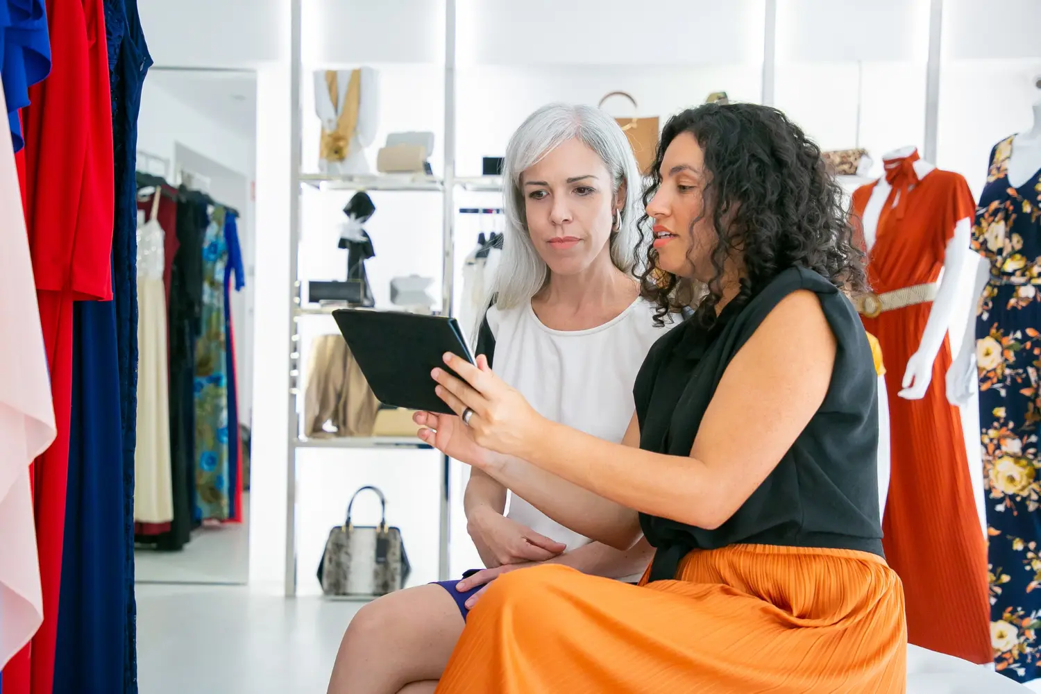 Two women looking at a tablet while browsing dresses in a boutique, symbolizing online search for where to watch VS fashion show.