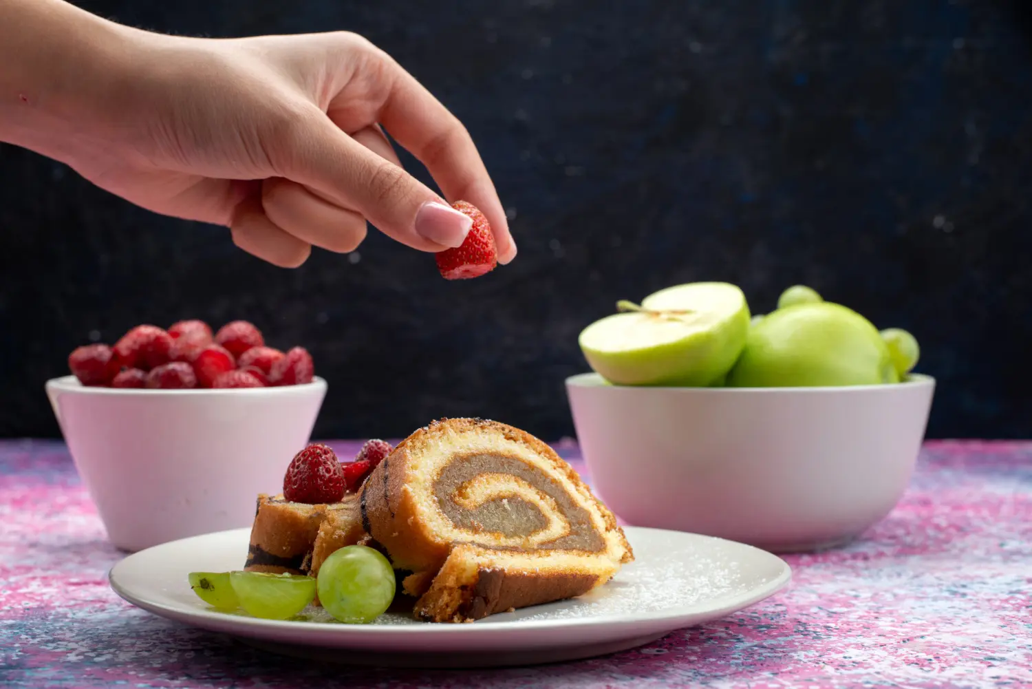 Hand placing a strawberry on a slice of rolled cake with grapes, with bowls of strawberries and green apples in the background.