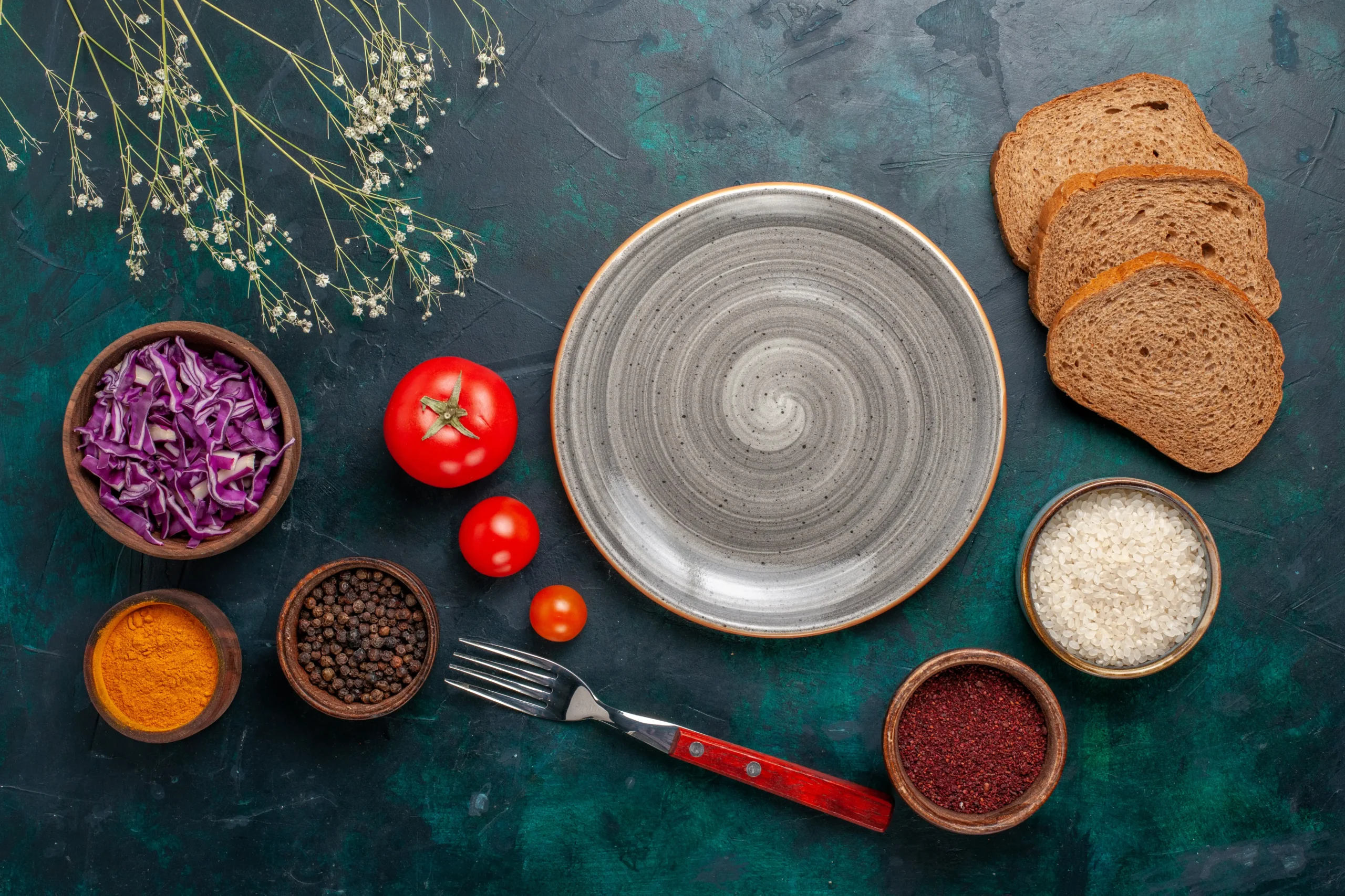 "Top view of an empty ceramic plate surrounded by tomatoes, sliced bread, rice, spices, purple cabbage, and a fork on a dark textured background."